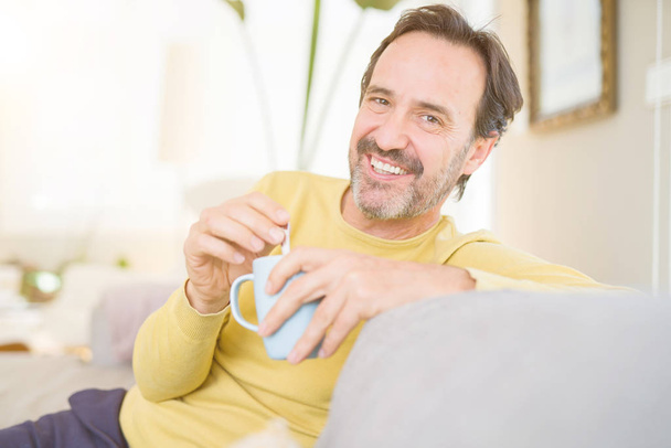 ハンサムな中年男がソファーに座ってリラックス コーヒーを一杯飲み、自宅のカメラで笑顔 - 写真・画像