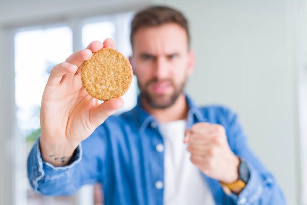 Hombre guapo comiendo saludable galleta de grano entero molesto y frustrado gritando con ira, loco y gritando con la mano levantada, concepto de ira
 - Foto, Imagen