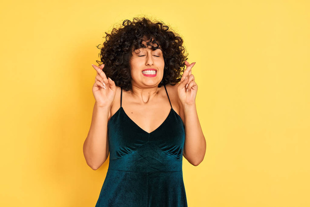 Young arab woman with curly hair wearing elegant dress over isolated yellow background gesturing finger crossed smiling with hope and eyes closed. Luck and superstitious concept. - Photo, Image
