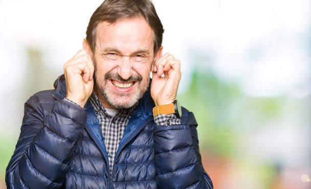 Średnim wieku przystojny mężczyzna na sobie płaszcz zimowy, obejmujące uszy palcami z zirytowany wyrażenie dla hałasu głośno muzyki. Koncepcja głuchy. - Zdjęcie, obraz