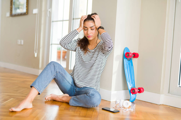 スケートボードとヘッドフォンを使って床に座っている若い女性は、頭痛に苦しんで、痛みや偏頭痛のために必死にストレスを受けました。頭の上に手を置く. - 写真・画像