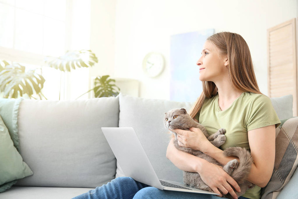 Beautiful woman with cute cat and laptop sitting on sofa at home - Fotó, kép