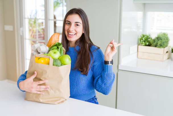 Красивая молодая женщина держит бумажный мешок, полный здоровых продуктов очень счастлив указывая рукой и пальцем в сторону
 - Фото, изображение