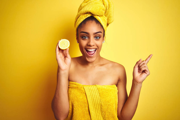 Afro kobieta nosi ręcznik po prysznicu trzymając plasterek cytryny na białym tle żółtym bardzo szczęśliwy wskazując ręką i palcem na bok - Zdjęcie, obraz