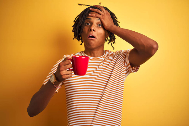 Afro american man with dreadlocks drinking a cup of coffee over isolated yellow background stressed with hand on head, shocked with shame and surprise face, angry and frustrated. Fear and upset for mistake. - Photo, image