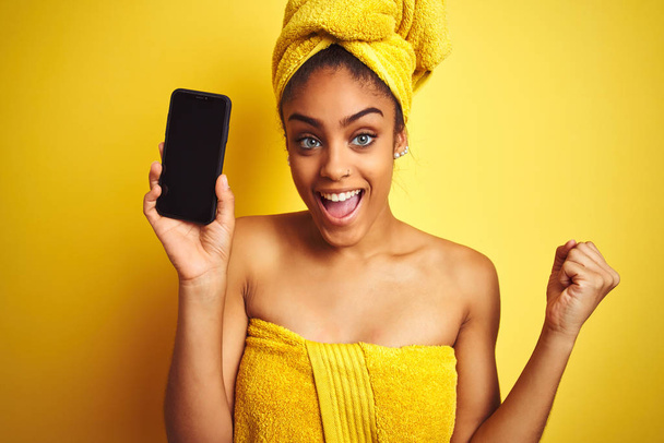 Afro femme portant serviette après douche montrant smatrphone sur fond jaune isolé criant fier et célébrant la victoire et le succès très excité, acclamant l'émotion
 - Photo, image