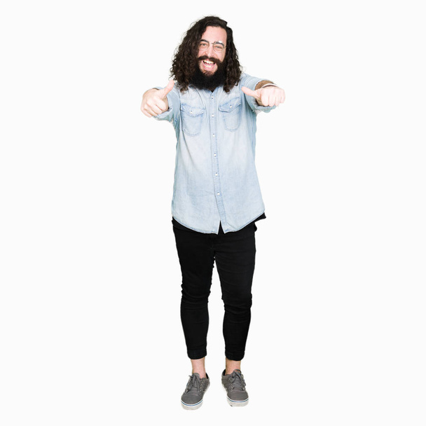 Young hipster man with long hair and beard wearing glasses approving doing positive gesture with hand, thumbs up smiling and happy for success. Looking at the camera, winner gesture. - Photo, Image