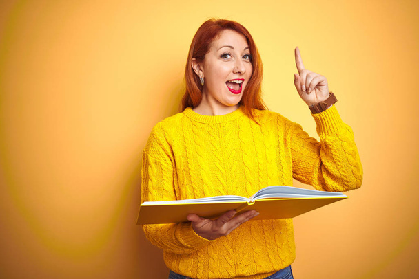 Νεαρή κοκκινομάλλα μαθήτρια που διαβάζει βιβλίο στέκεται πάνω από το κίτρινο απομονωμένο φόντο έκπληκτος με μια ιδέα ή ερώτηση που δείχνει το δάχτυλο με χαρούμενο πρόσωπο, νούμερο ένα - Φωτογραφία, εικόνα