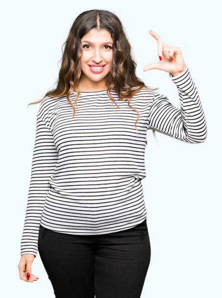 junge schöne Frau in Streifen-Pullover lächelnd und selbstbewusst gestikulierend mit der Hand, die mit den Fingern ein Zeichen setzt, während sie in die Kamera schaut. Maßnahmenkonzept. - Foto, Bild