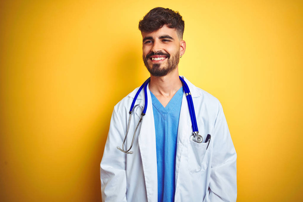 Молодий лікар з татуюванням, одягнений у стетоскоп, стоїть над ізольованим жовтим тлом, дивлячись на бік з посмішкою на обличчі, природним виразом. Сміється впевнено
. - Фото, зображення