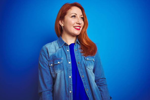 Young beautiful redhead woman wearing denim shirt standing over blue isolated background looking away to side with smile on face, natural expression. Laughing confident. - Photo, Image
