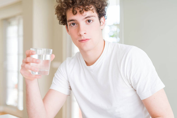 Jovem bebendo um copo de água em casa com uma expressão confiante no rosto inteligente pensando sério
 - Foto, Imagem