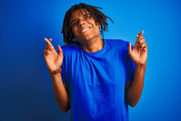 Афро-американец с дредами в футболке, стоящей на изолированном синем фоне, жестом скрестив пальцы, улыбаясь с надеждой и закрыв глаза. Удача и суеверие
. - Фото, изображение