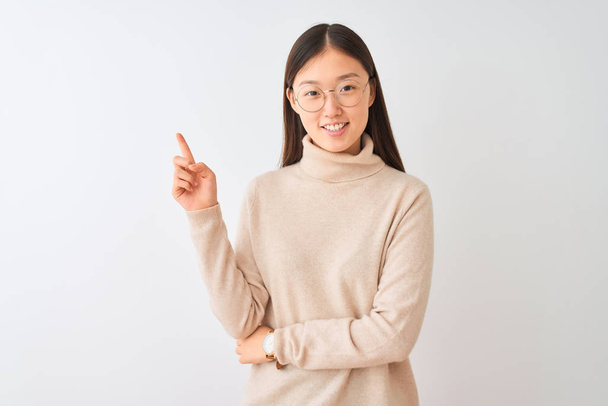 Fiatal kínai nő viselt garbó pulóvert és szemüveg alatt elszigetelt fehér háttér egy nagy mosollyal az arcon, mutatva a kezét, és ujját az oldalon nézi a kamerát. - Fotó, kép