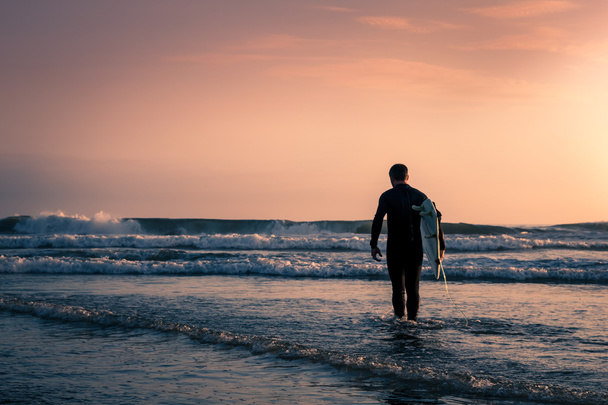 Homem surfista de fato de mergulho preto com prancha branca na praia ao pôr-do-sol
. - Foto, Imagem