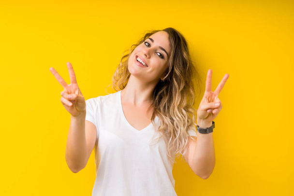 Young beautiful blonde woman over yellow background smiling looking to the camera showing fingers doing victory sign. Number two. - Foto, immagini
