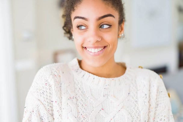 Mulher americana africana bonita jovem com cabelo afro vestindo camisola casual sorrindo lado olhando e olhando para longe pensando
. - Foto, Imagem