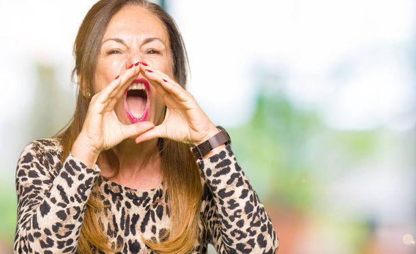 Όμορφη μέση ηλικία γυναίκα φοράει λεοπάρδαλη ζώο print φόρεμα φωνάζει θυμωμένος δυνατά με τα χέρια πάνω από το στόμα - Φωτογραφία, εικόνα