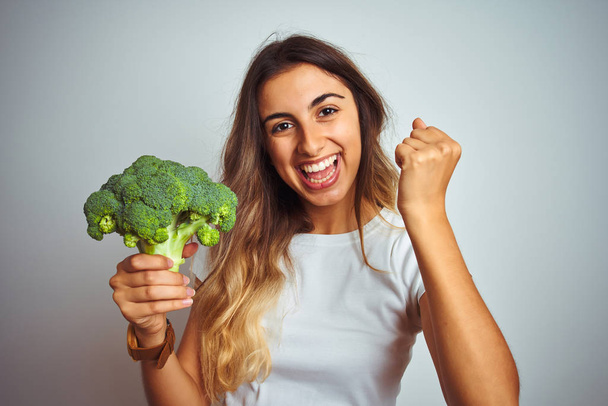 Young beautiful woman eating broccoli over grey isolated background screaming proud and celebrating victory and success very excited, cheering emotion - Foto, imagen