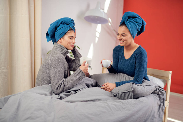 Dos hermosas amigas caucásicas con toallas en la cabeza sentadas en la cama en el dormitorio, bebiendo café y chismorreando
. - Foto, Imagen