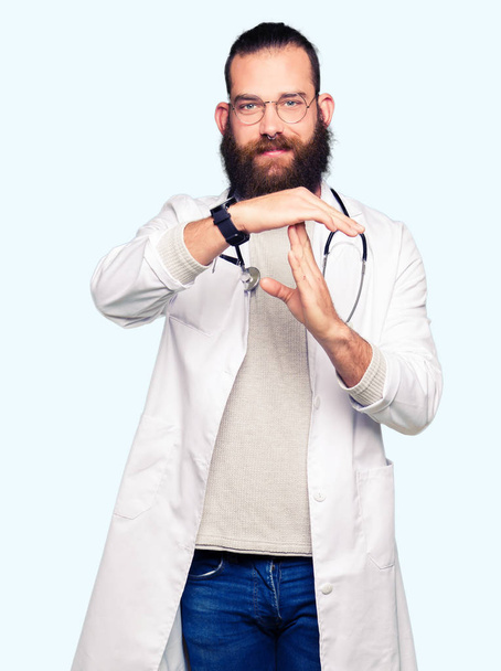 Молодой блондин-врач с бородой, в медицинском пальто, делает тайм-аут жестом руками, расстроенным и серьезным лицом
 - Фото, изображение