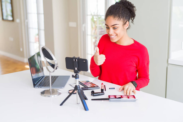 Young african american influencer woman recording make up tutorial doing happy thumbs up gesture with hand. Approving expression looking at the camera with showing success. - Photo, Image
