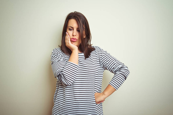 Young beautiful brunette woman wearing striped sweater over white isolated background thinking looking tired and bored with depression problems with crossed arms. - 写真・画像
