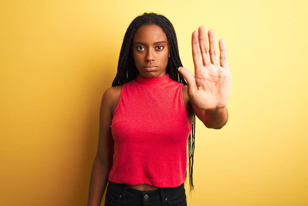 Mujer afroamericana vistiendo camiseta casual roja de pie sobre fondo amarillo aislado haciendo dejar de cantar con la palma de la mano. Expresión de advertencia con gesto negativo y serio en la cara
. - Foto, Imagen