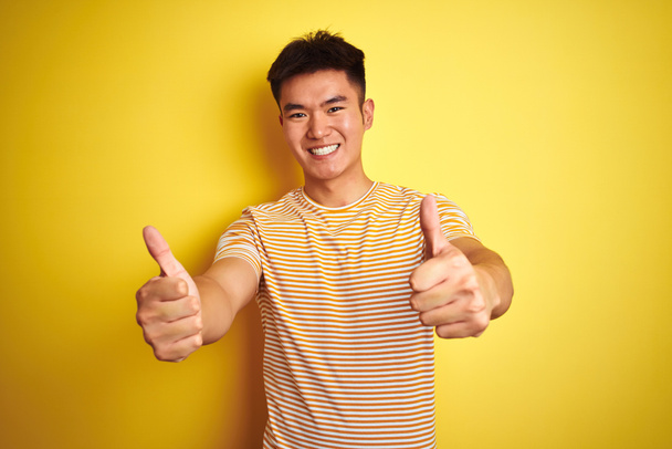Young asian chinese man wearing t-shirt standing over isolated yellow background approving doing positive gesture with hand, thumbs up smiling and happy for success. Winner gesture. - Фото, зображення