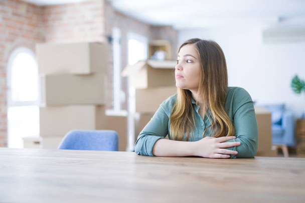 Young woman sitting on the table with cardboard boxes behind her moving to new home looking to side, relax profile pose with natural face with confident smile. - Photo, image