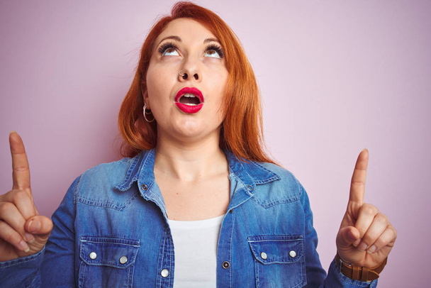 Youg beautiful redhead woman wearing denim shirt standing over isolated pink background amazed and surprised looking up and pointing with fingers and raised arms. - Photo, Image