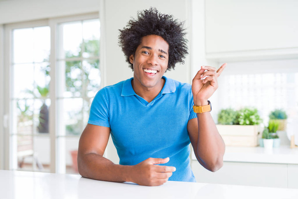 African American man thuis met een grote glimlach op het gezicht, met de hand en de vinger te wijzen naar de kant te kijken naar de camera. - Foto, afbeelding