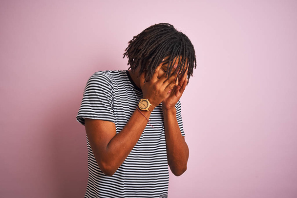 ネイビーのストライプTシャツを着たドレッドロックを着たアフロ男は、泣きながら手で顔を覆う悲しい表情で孤立したピンクの背景の上に立っています。うつ病の概念. - 写真・画像