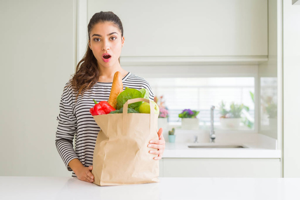 Young woman holding paper bag full of groceries scared in shock with a surprise face, afraid and excited with fear expression - Foto, imagen
