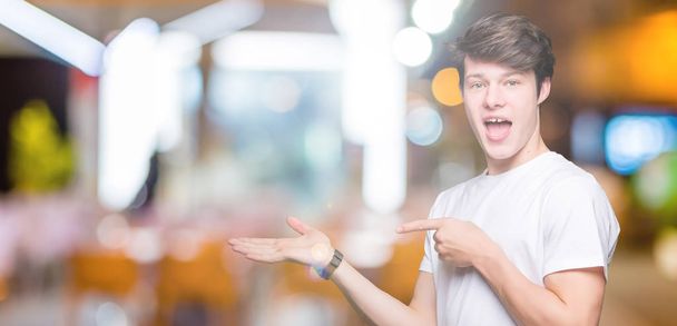 Jonge knappe man dragen casual wit t-shirt over geïsoleerde achtergrond verbaasd en lachend naar de camera tijdens het presenteren met hand en met de vinger te wijzen. - Foto, afbeelding
