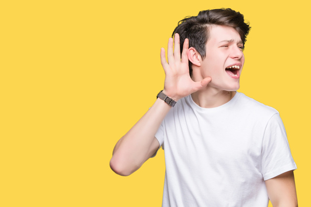 junger gutaussehender Mann in lässigem weißem T-Shirt vor isoliertem Hintergrund, der lächelnd mit der Hand über dem Ohr Gerüchten oder Klatsch lauscht. Konzept für Gehörlosigkeit. - Foto, Bild