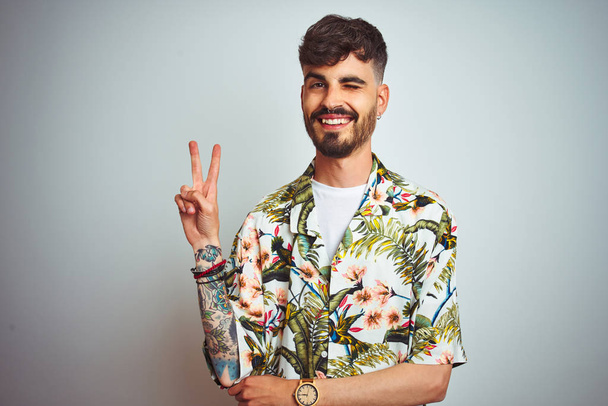 Чоловік з татуюванням на канікулах у літній сорочці стоїть над ізольованим білим тлом, посміхаючись із щасливим обличчям, підморгнутим до камери, роблячи знак перемоги. Номер два
. - Фото, зображення