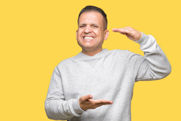 Hombre árabe de mediana edad con sudadera deportiva sobre fondo aislado gesticulando con las manos mostrando un signo de tamaño grande y grande, símbolo de medida. Sonriendo mirando a la cámara. Concepto de medición
. - Foto, Imagen