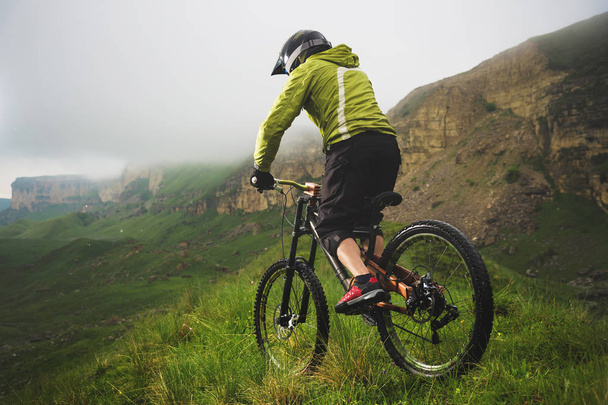 Aged mannelijke atleet in helm en masker rijdt een mountainbike op een grasachtige helling tegen de achtergrond van plateau rotsen en lage wolken op een bewolkte dag. Downhill mountainbike concept - Foto, afbeelding