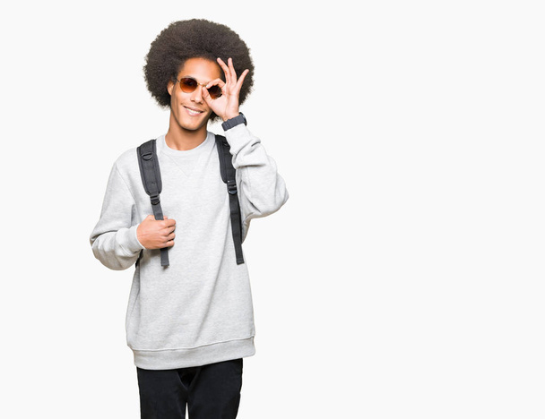 Joven hombre afroamericano con pelo afro usando gafas de sol y mochila con la cara feliz sonriendo haciendo signo bien con la mano en el ojo mirando a través de los dedos
 - Foto, imagen