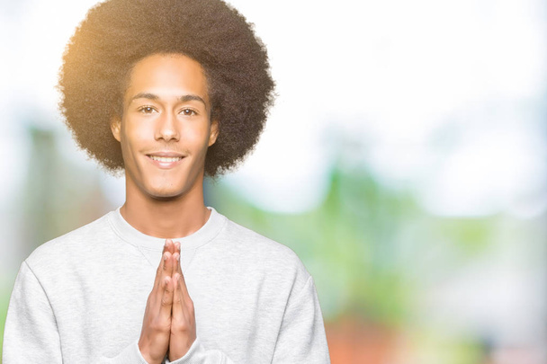 Joven afroamericano con cabello afro usando sudadera deportiva rezando con las manos juntas pidiendo perdón sonriendo confiado
. - Foto, Imagen