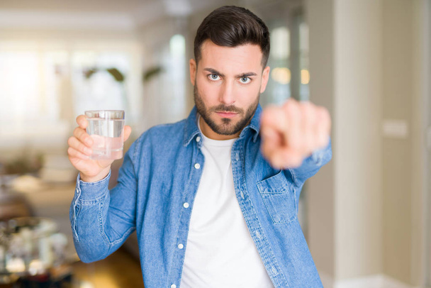 Όμορφος νεαρός πίνει ένα ποτήρι νερό στο σπίτι δείχνοντας με το δάχτυλο στη φωτογραφική μηχανή και σε εσάς, χέρι σημάδι, θετική και σίγουρη κίνηση από το μέτωπο - Φωτογραφία, εικόνα