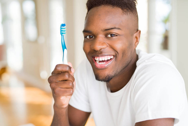 Африканский американец держит зубную щетку со счастливым лицом стоя и улыбаясь с уверенной улыбкой, показывая зубы
 - Фото, изображение