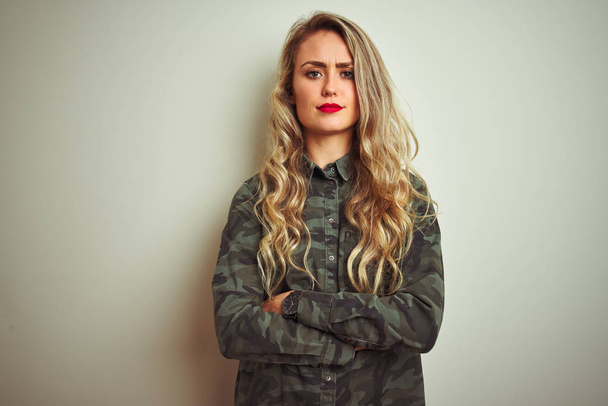 Jonge mooie vrouw dragen militaire camouflage shirt over witte geïsoleerde achtergrond scepticus en nerveus, afkeurend uitdrukking op gezicht met gekruiste armen. Negatieve persoon. - Foto, afbeelding