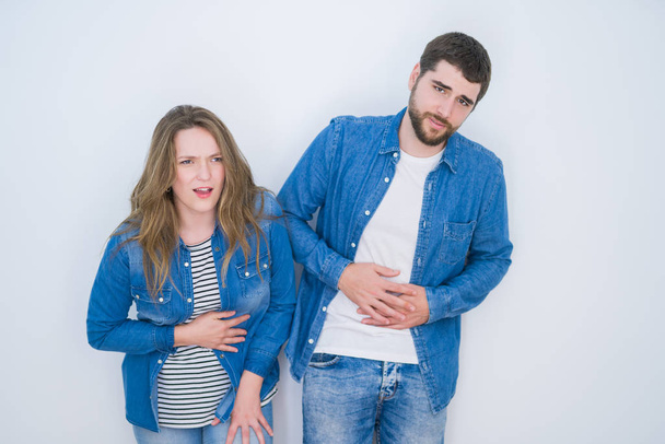 Young beautiful couple standing together over white isolated background with hand on stomach because indigestion, painful illness feeling unwell. Ache concept. - Foto, Imagen