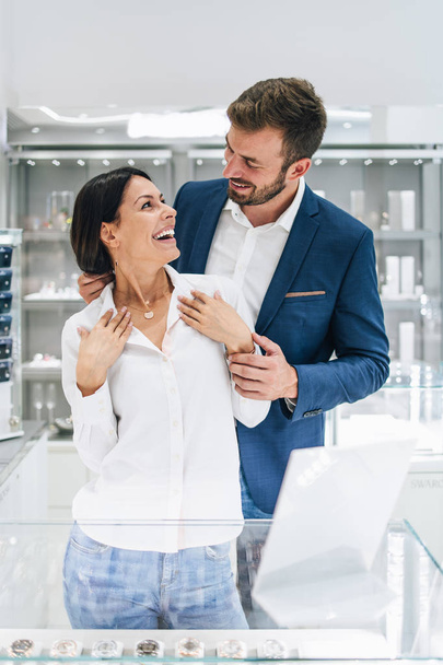 Beautiful couple enjoying in shopping at modern jewelry store. Young woman try it out gorgeous necklace and earrings. - Φωτογραφία, εικόνα