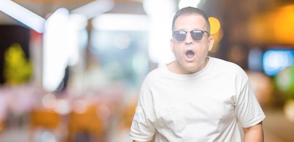 Arabský muž středního věku s bílým tričkem a slunečními brýlemi na izolovaném pozadí se bojí a šokuje překvapením, strachem a vzrušovaným obličejem. - Fotografie, Obrázek