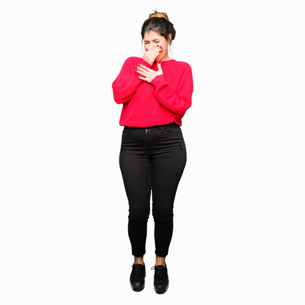 Young beautiful woman wearing red sweater and bun smelling something stinky and disgusting, intolerable smell, holding breath with fingers on nose. Bad smells concept. - Photo, image
