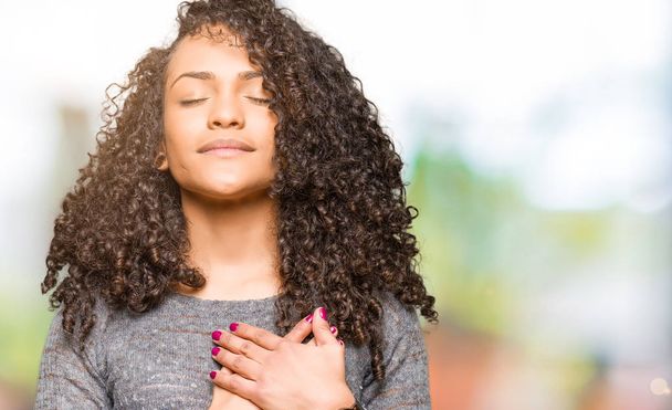 Young beautiful woman with curly hair wearing grey sweater smiling with hands on chest with closed eyes and grateful gesture on face. Health concept. - Foto, Imagen
