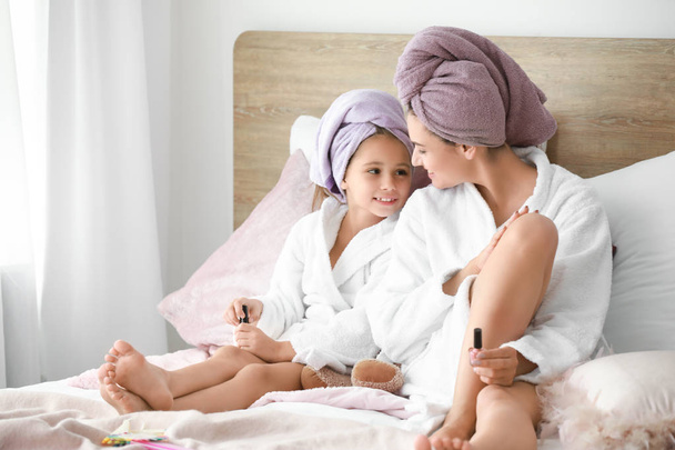 Moeder en haar kleine dochter in badjassen doen pedicure in slaapkamer - Foto, afbeelding